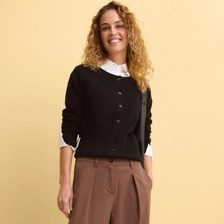 Jachetă tricotată neagră cu nasturi peste o bluză albă, combinată cu pantaloni maro.