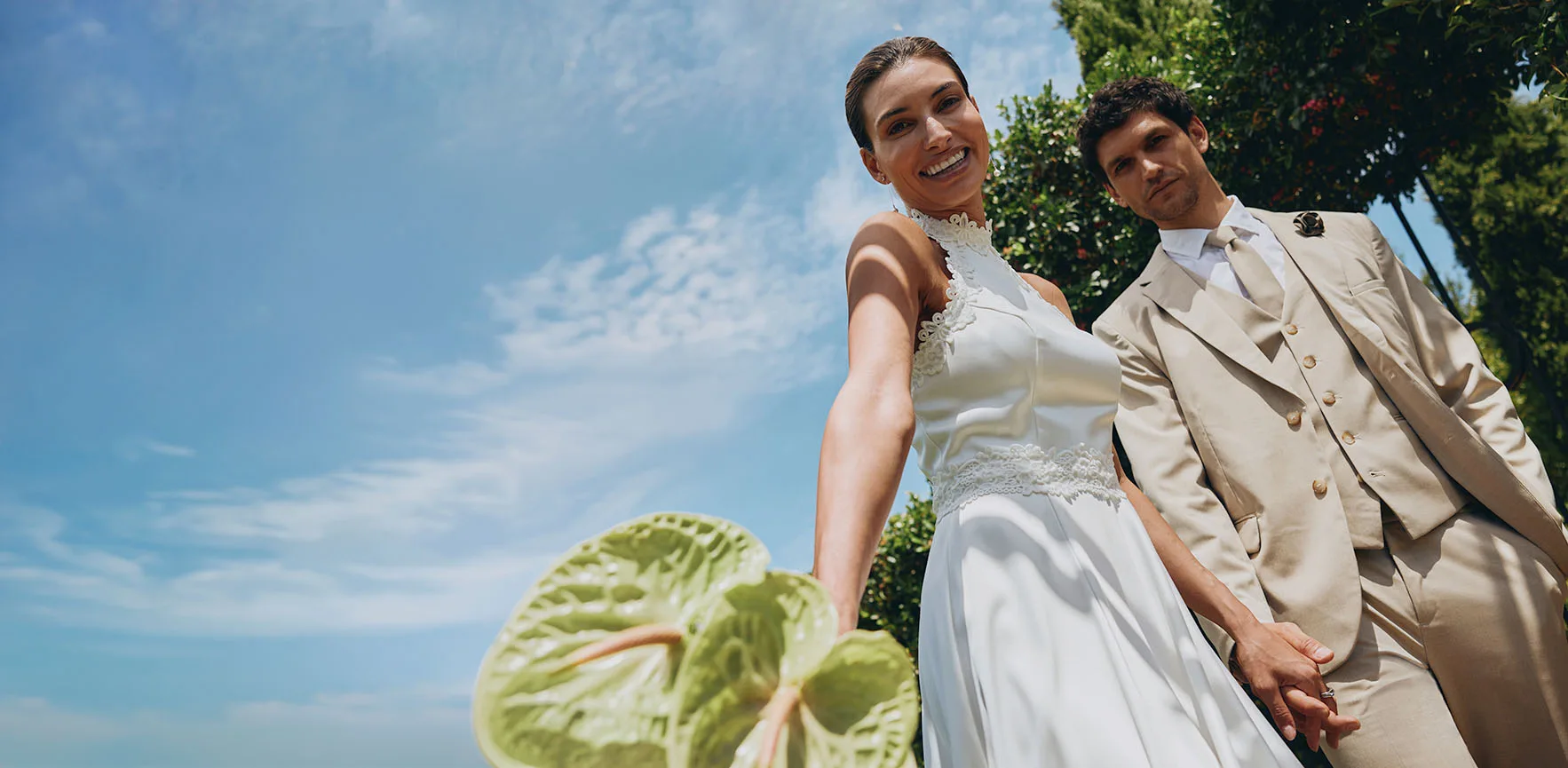 Pereche elegantă de miri scăldați în razele soarelui. Ea în rochie albă, el în constum bej cu ochelari de soare.