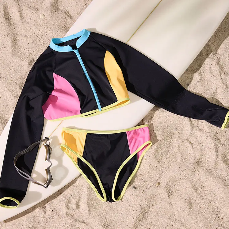Costum de baie colorat cu mânecă lungă și bikini pe nisip lângă o placă de surf.