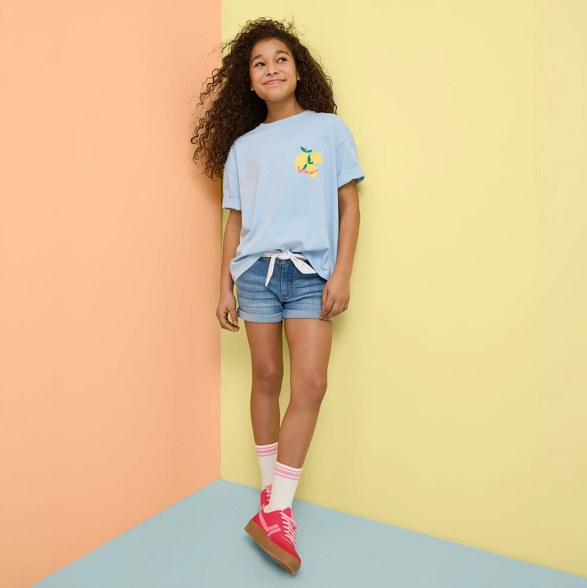Ținută de vară cu tricou bleu, pantaloni scurți de blugi și pantofi sport colorați.
