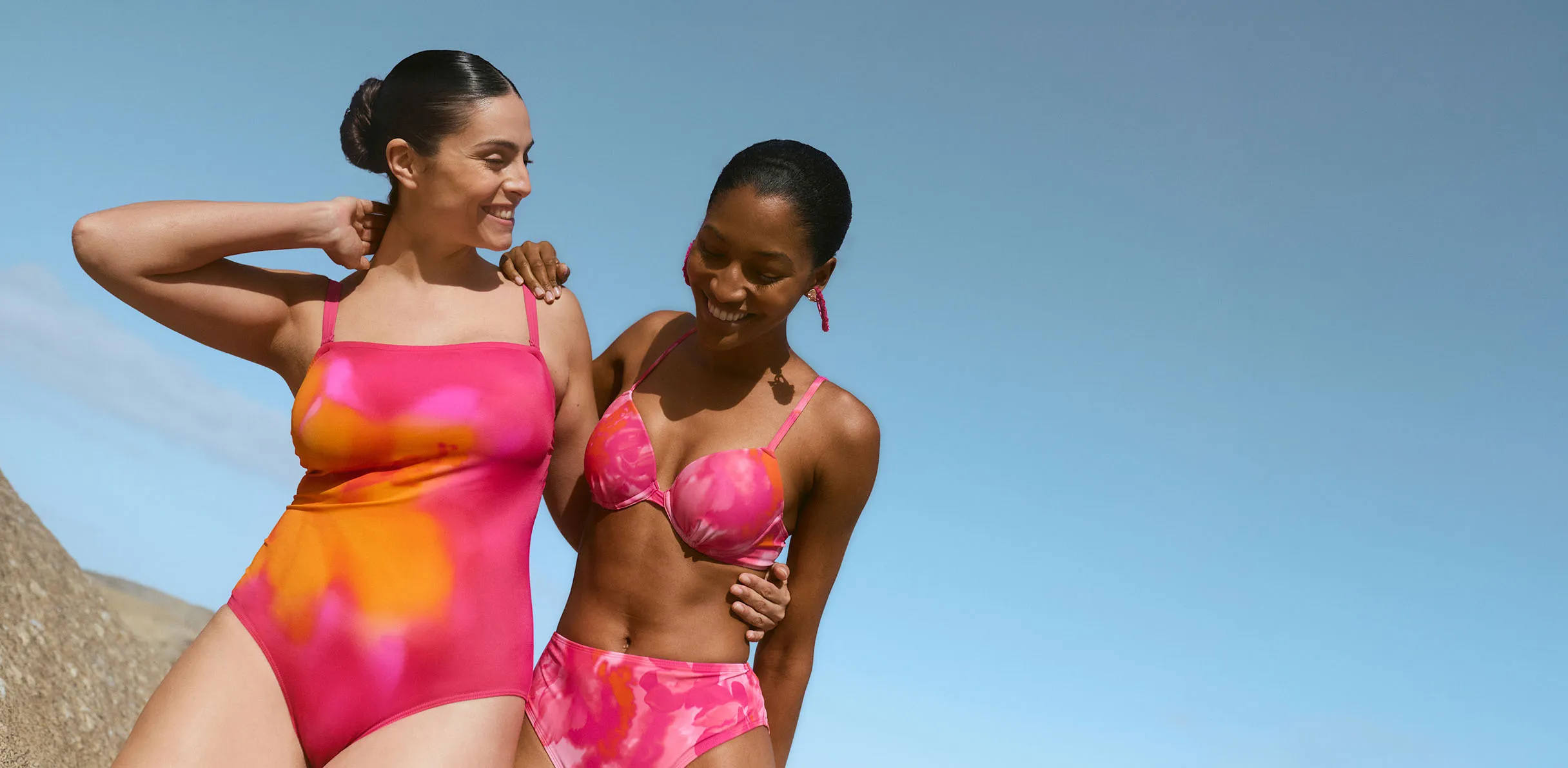 Două femei cu costum de baie bikini și costum de baie întreg pink fluorescent, la plajă.