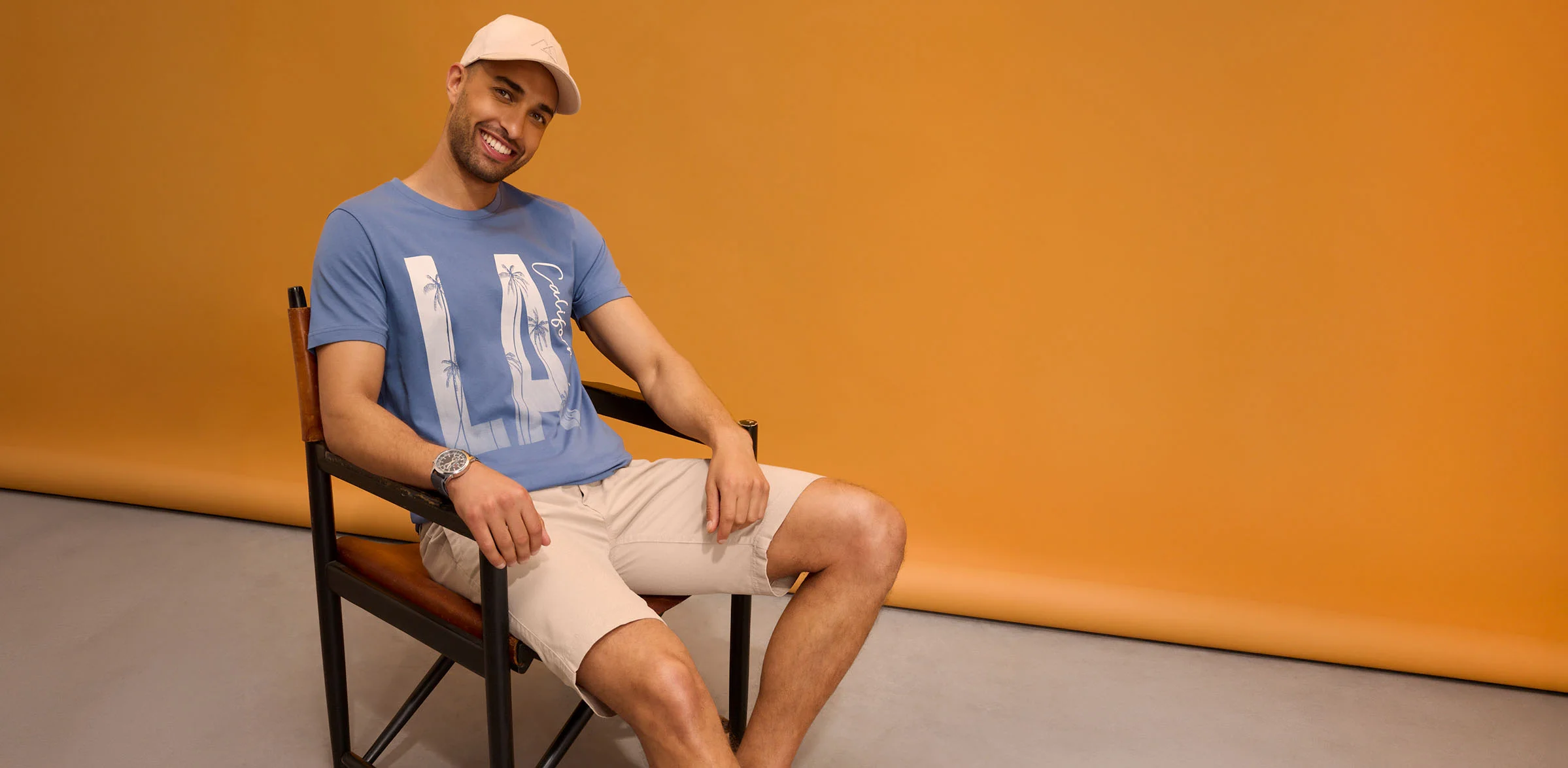 O ținută casual: șapcă, tricou albastru cu imprimeu, pantaloni bej scurți și ceas de mână.